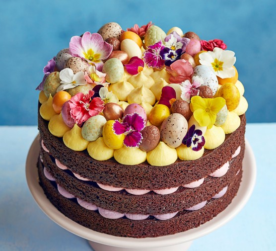 Layered chocolate cake with yellow icing topped with edible flowers and mini eggs